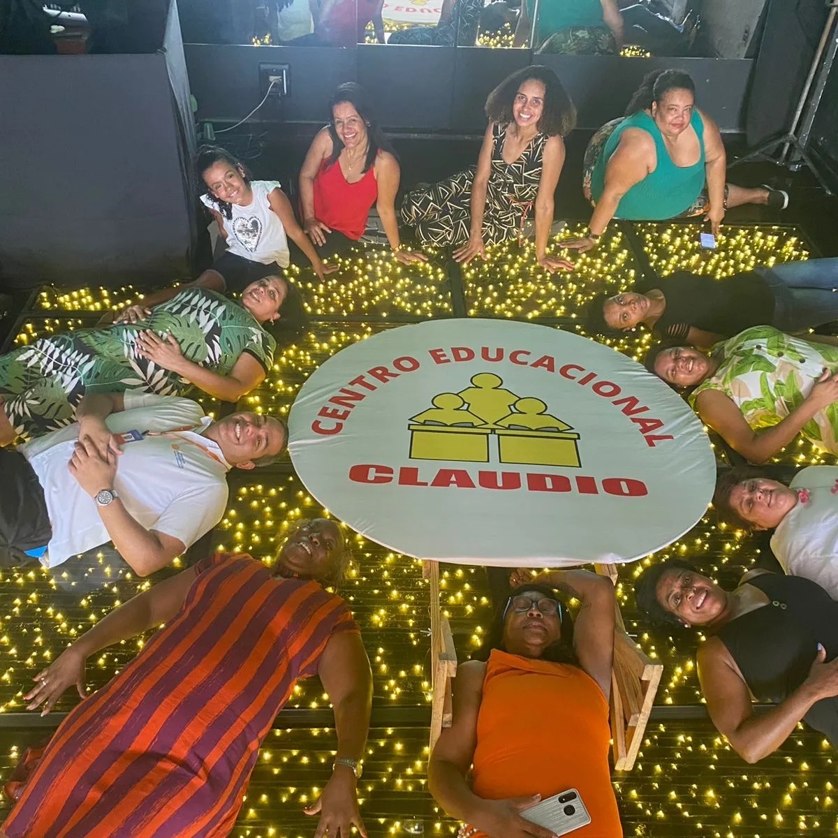Equipe do Centro Educacional Claudio deitada em roda, sorrindo, ao redor de uma mesa com o logo da escola