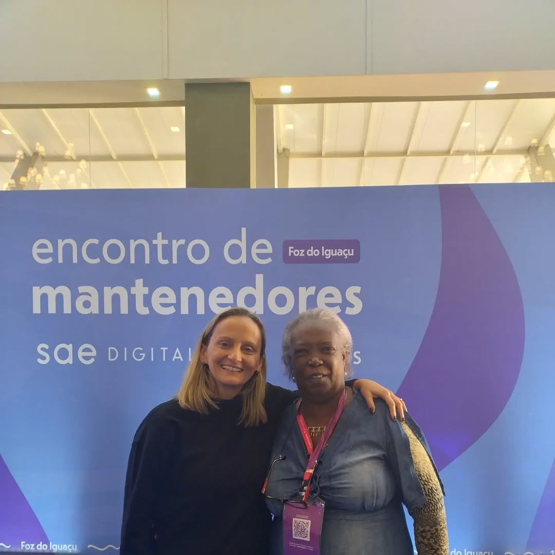 Duas mulheres sorrindo diante do painel do Encontro de Mantenedores do SAE Digital em Foz do Iguaçu